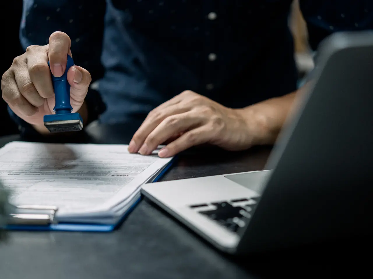 A person stamps a document on a clipboard while sitting at a desk with an open laptop. Only their hands and torso are visible.