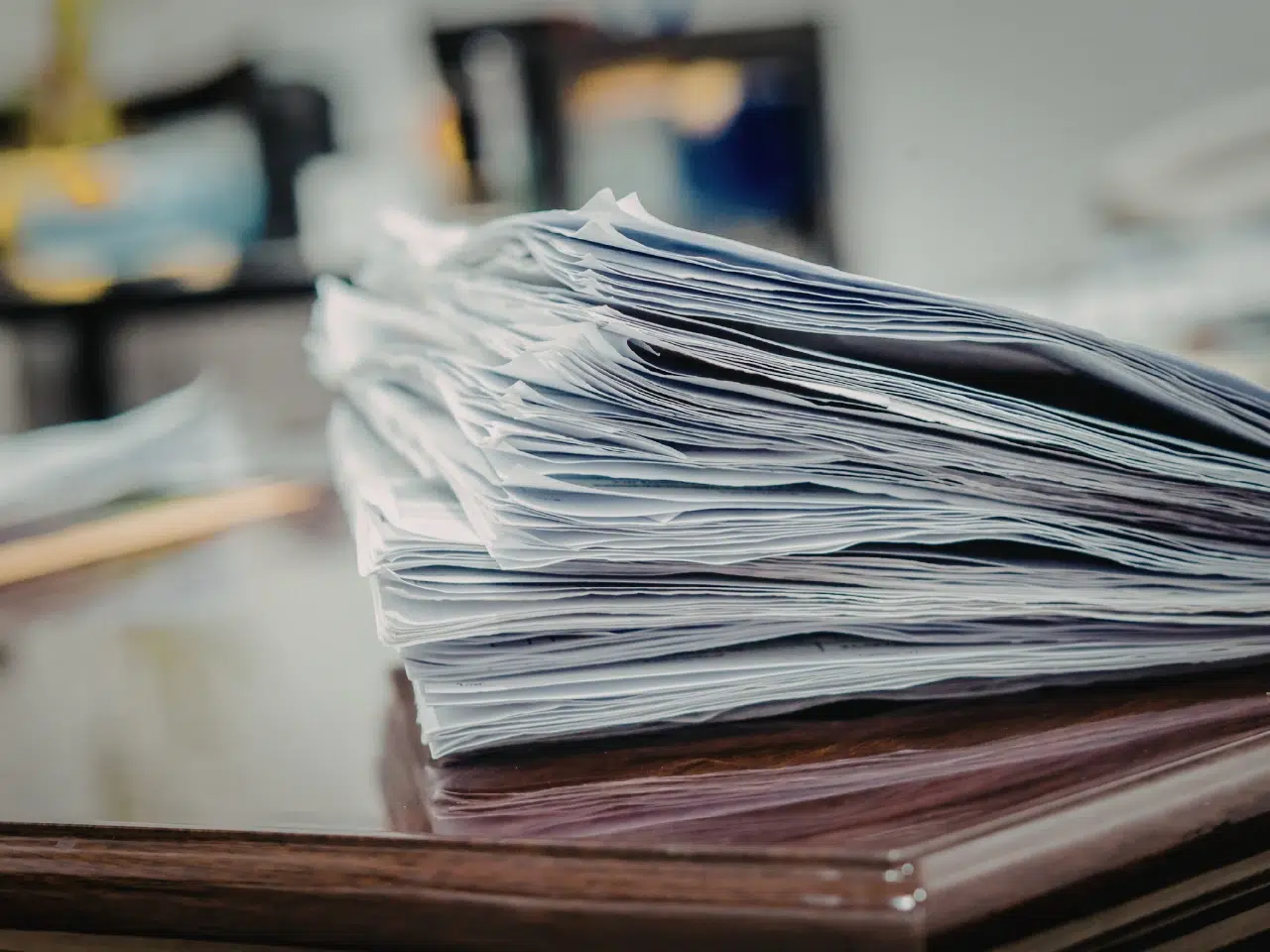 A large stack of papers is piled on a wooden desk, with some sheets appearing slightly crumpled. The background is out of focus, suggesting an office or workspace environment.