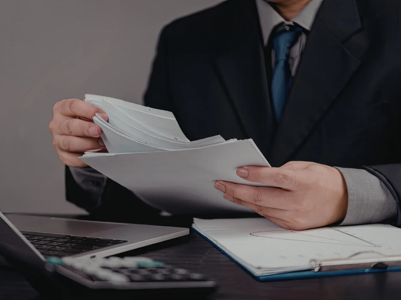 A person in a suit sits at a desk, reviewing a stack of documents. An open laptop, calculator, and a notepad are on the desk, suggesting a professional or office setting.