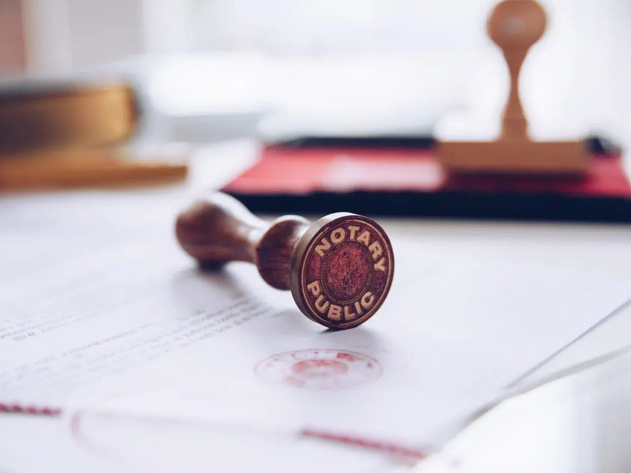 A notary public stamp rests on a document, with another stamp and office supplies blurred in the background. The focus is on the stamp’s engraved seal facing the camera.