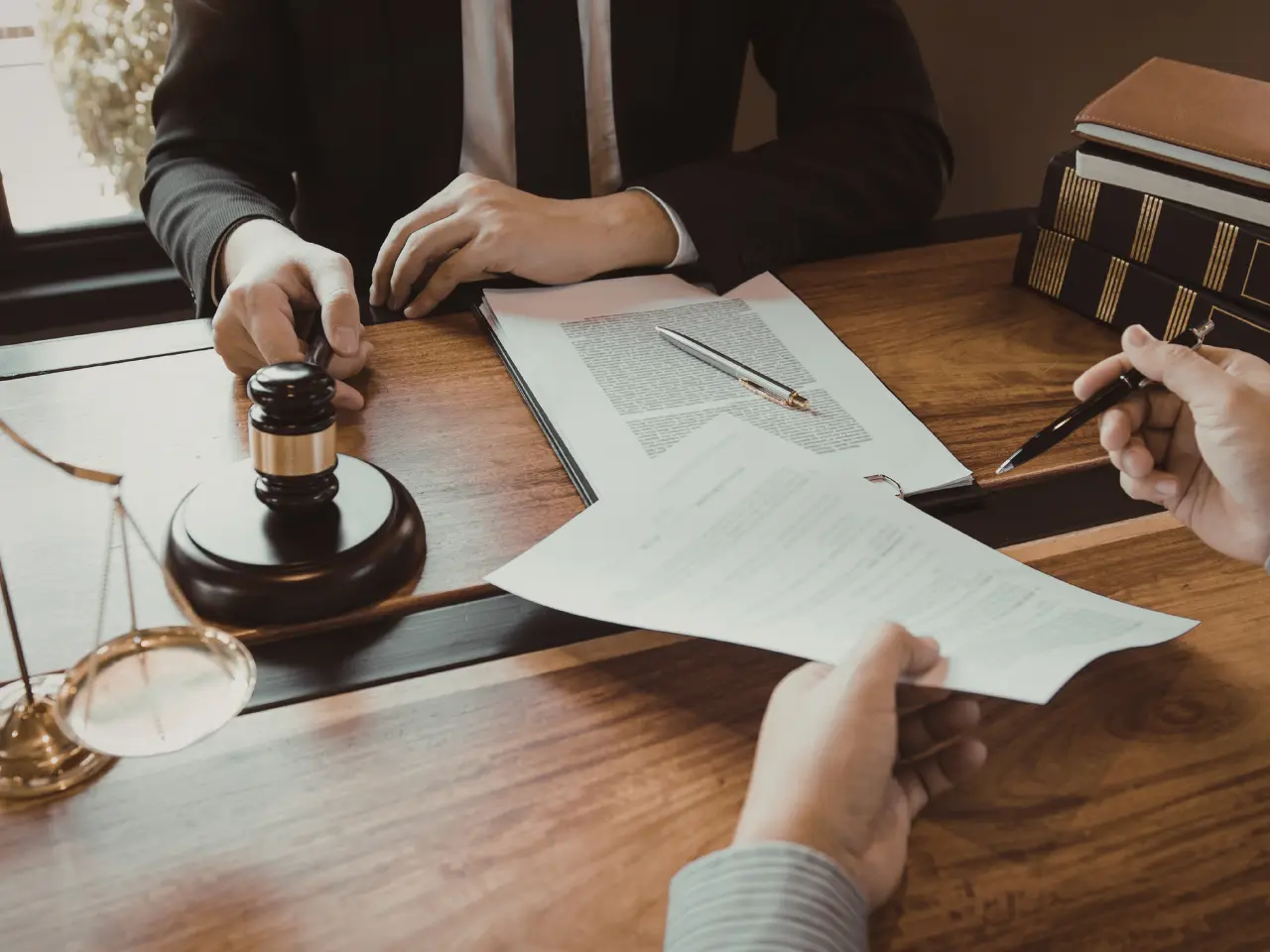 Two people sit at a wooden desk with legal documents, a gavel, and scales of justice. One person is signing a paper while the other, possibly a lawyer, gestures with their hand.