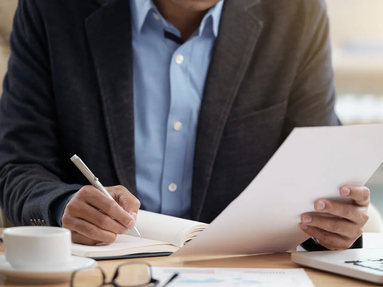 A person in a suit jacket writes in a notebook while holding a sheet of paper at a desk with a laptop, glasses, and a cup of coffee. The face is not visible.