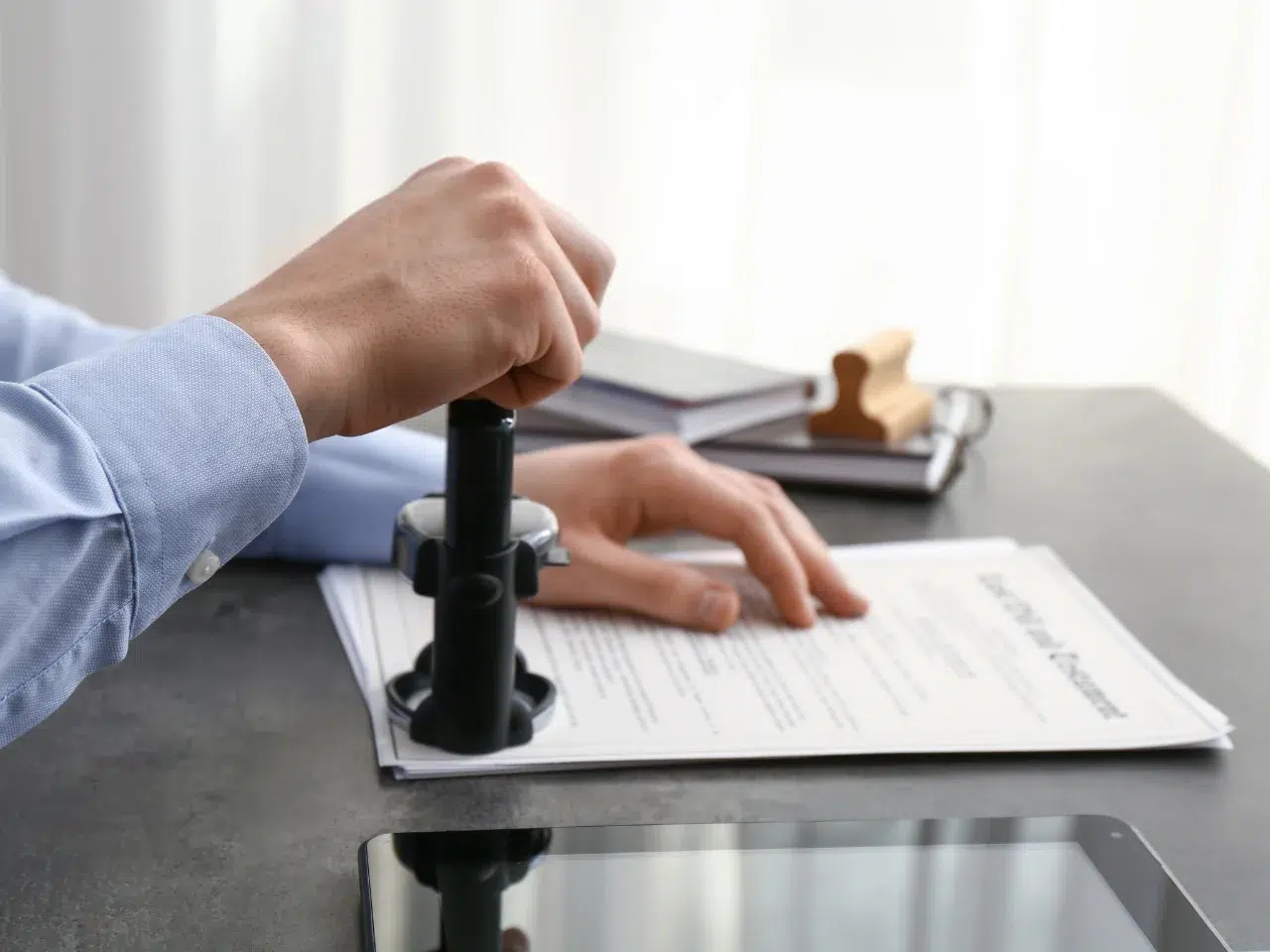 A person in a blue shirt stamps a stack of documents on a desk, with a notary seal visible. Other office supplies, including books and a tablet, are also on the desk.