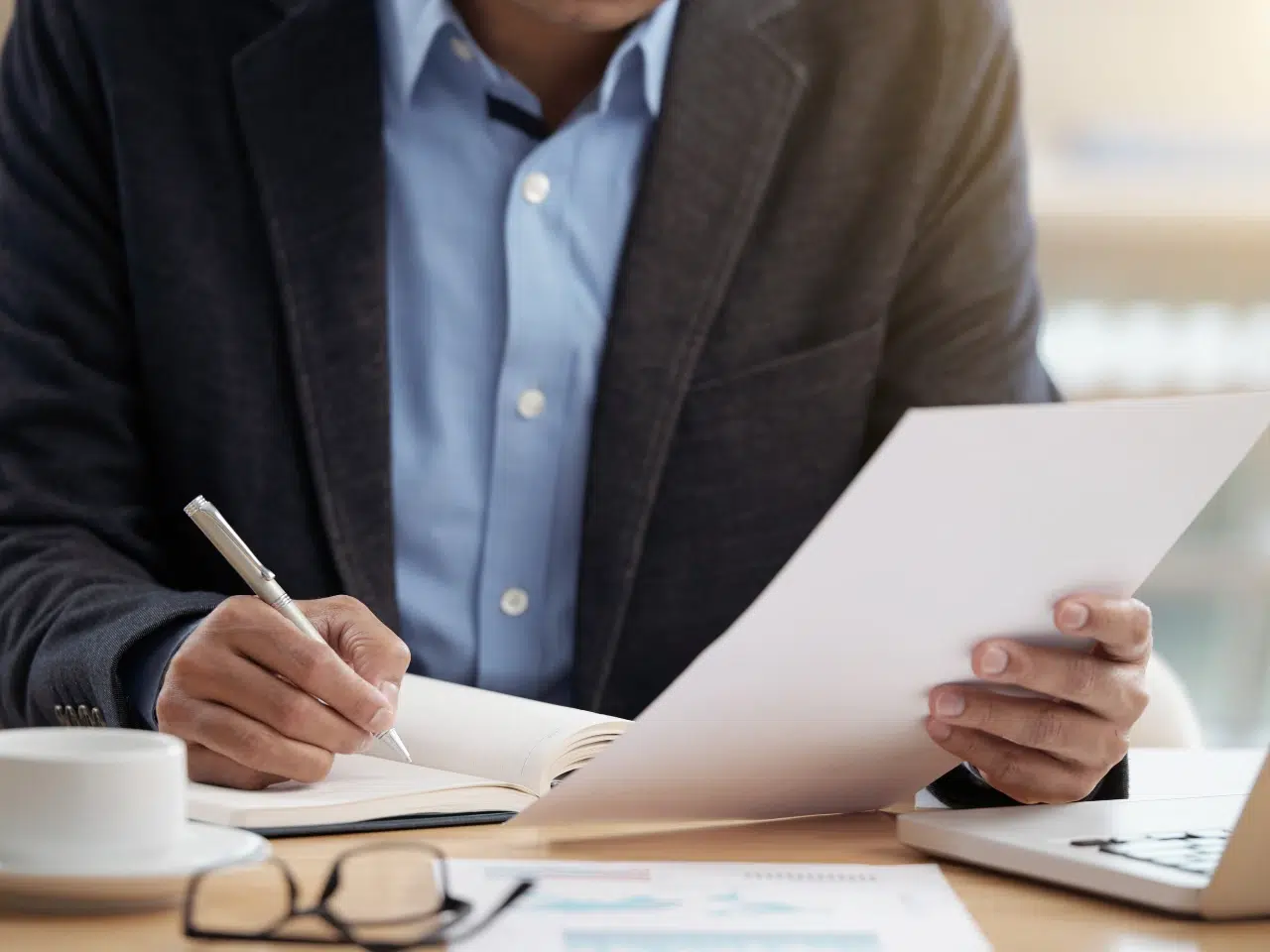 A person in a suit jacket and blue shirt sits at a desk, holding a sheet of paper in one hand and writing in a notebook with the other. Nearby are eyeglasses, documents, a laptop, and a cup of coffee.