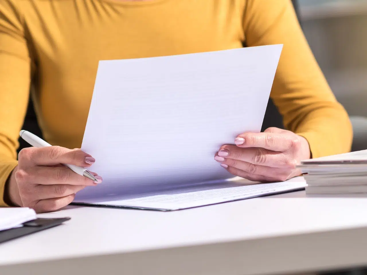 A person wearing a yellow top sits at a desk, holding and reading a document with both hands. A pen and a stack of papers are also visible on the desk.
