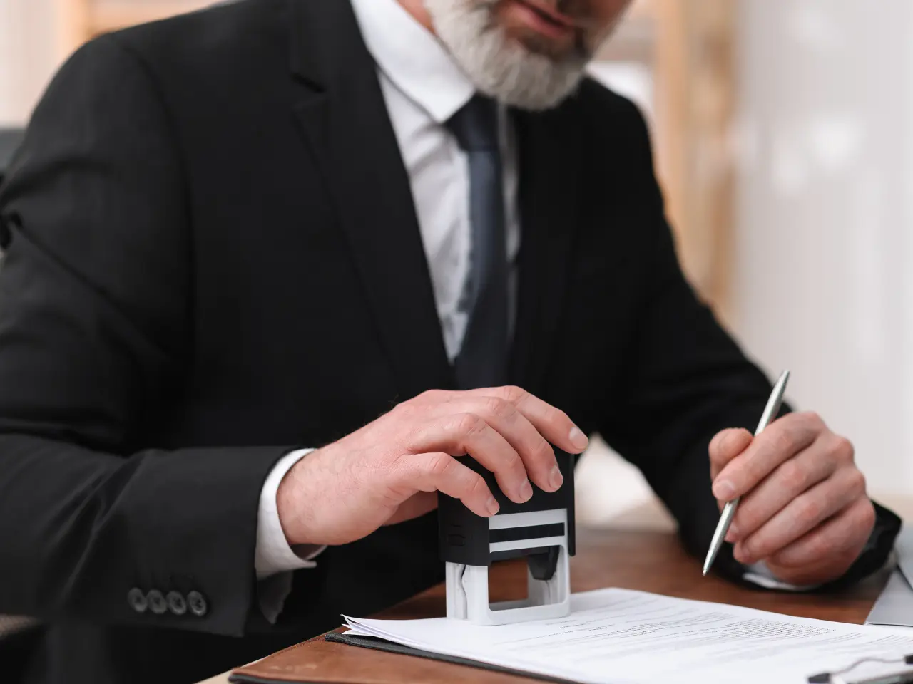 A man in a suit presses a stamp onto a document on a desk, holding a pen in his other hand. The focus is on his hands and the paperwork, suggesting an official or legal process.
