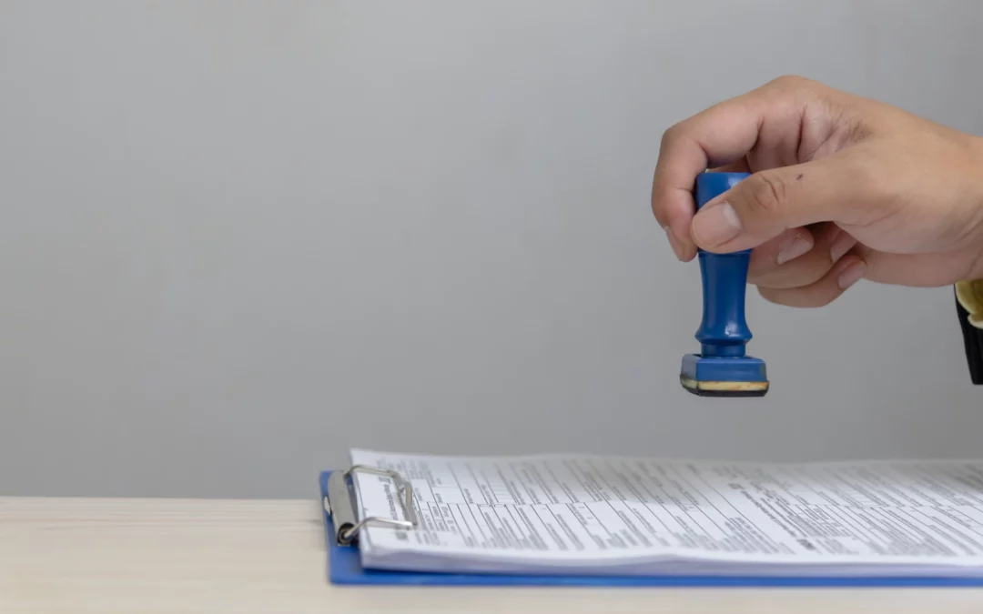 A hand holding a blue rubber stamp is about to stamp a document attached to a blue clipboard on a light-colored desk.