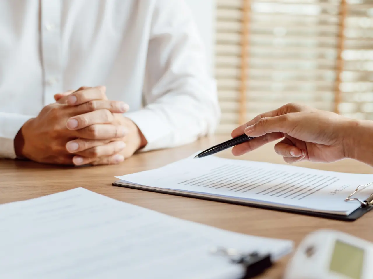 Two people sit at a desk; one has hands clasped, while the other holds a pen and gestures toward a document on the table, suggesting a discussion or business meeting.