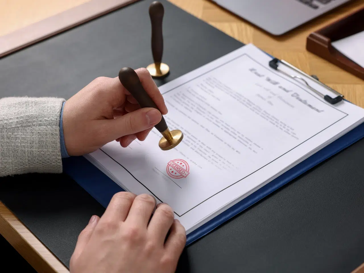 A person stamps a document attached to a clipboard using a wooden-handled stamp. Another stamp rests nearby on the desk, with part of a laptop and a tray visible in the background.