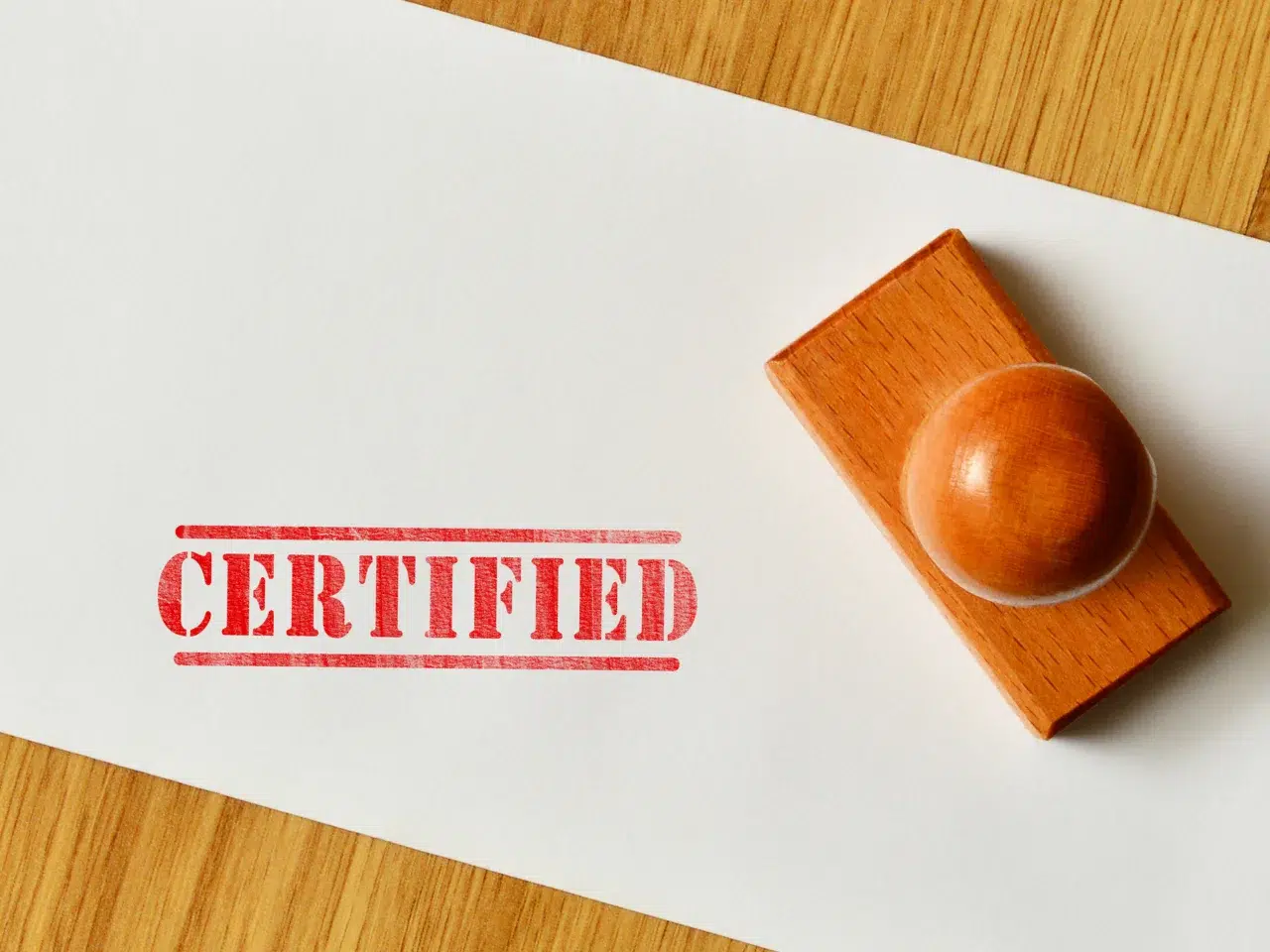 A wooden stamp rests on a white sheet of paper with the word CERTIFIED stamped in bold red letters. The paper is on a wooden surface.