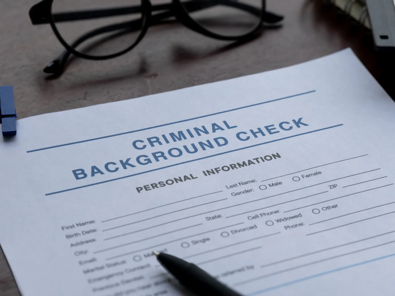 A close-up of a criminal background check form with sections for personal information. A black pen rests on the paper, and eyeglasses are visible in the background on a wooden surface.