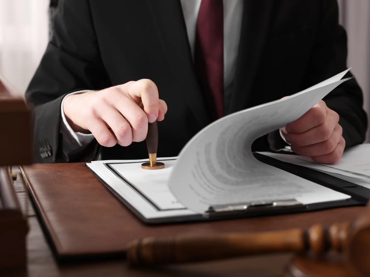 A person in a black suit stamps a document on a desk, with a wooden gavel and legal files visible nearby.