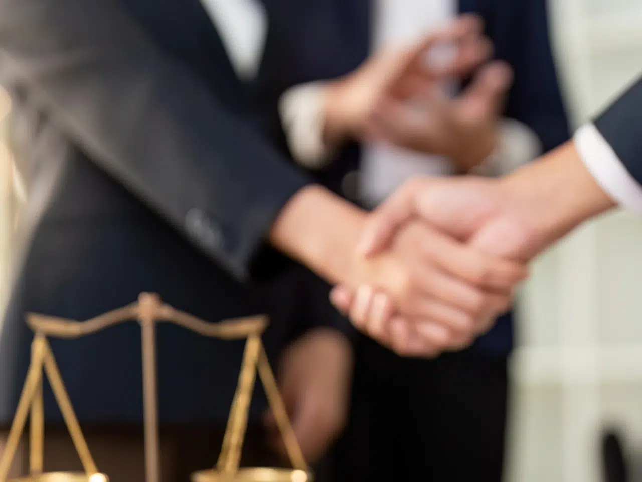 Two people in business attire shaking hands, with a blurred set of golden scales in the foreground and more people in the background, suggesting a professional or legal agreement.