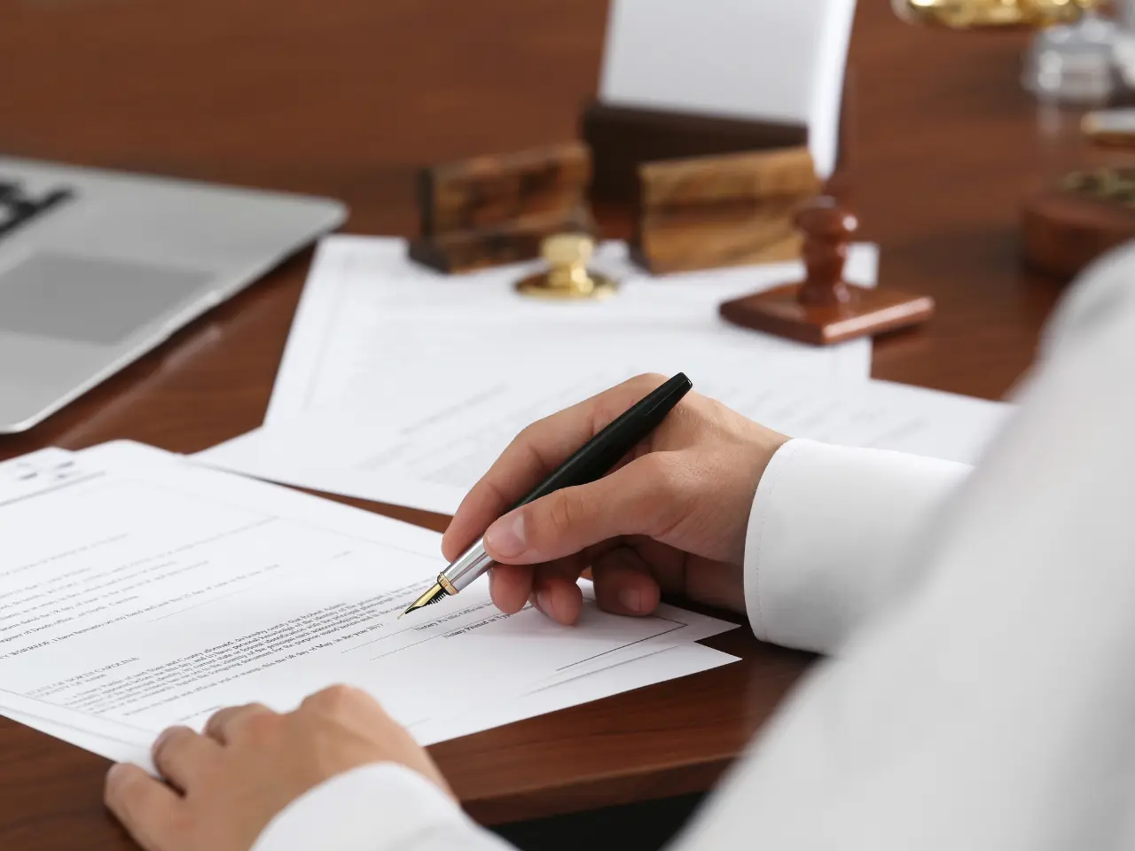 A person in a white shirt is signing a document with a pen at a desk, surrounded by paperwork, a laptop, and wooden stamps.