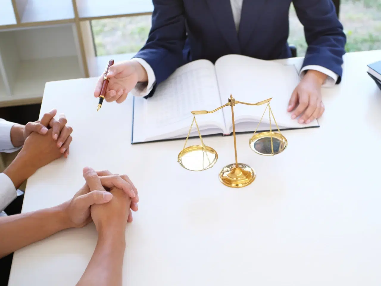 Three people sit at a table with a legal scale in the center. One person in a suit gestures with a pen over an open book, while the other two sit with hands clasped, suggesting a legal consultation.