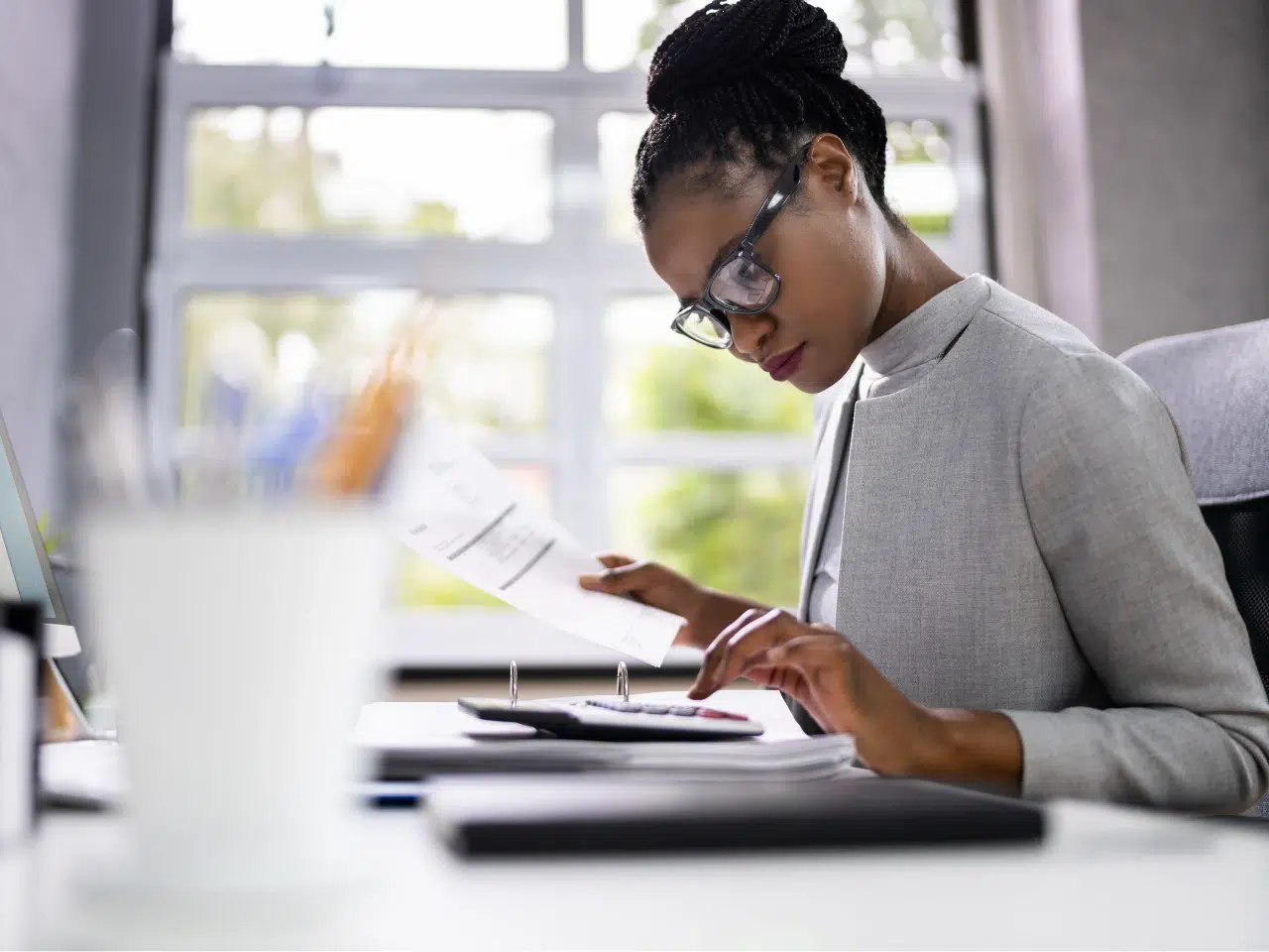 A woman wearing glasses sits at a desk, holding papers in one hand while using a calculator with the other. She appears focused, with office supplies and a window in the background.