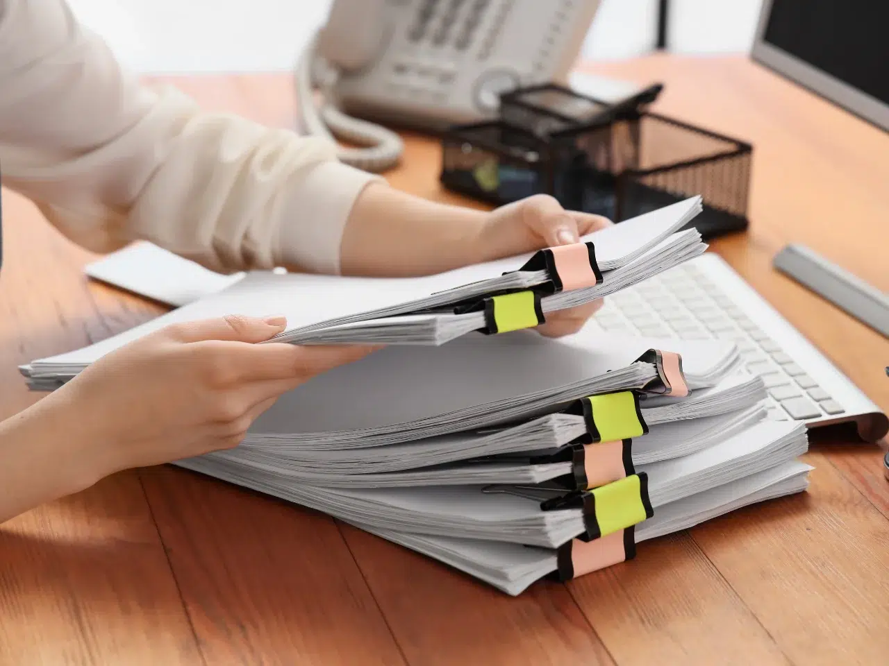 A person organizing several large stacks of documents clipped with binder clips on a wooden desk, with a keyboard, office phone, and wire organizers in the background.