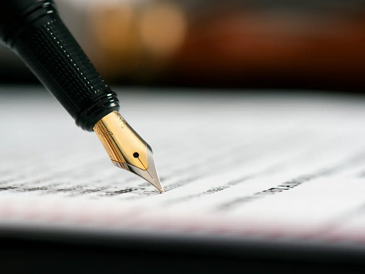 A close-up of a gold and black fountain pen writing on a sheet of paper with blurred text, focusing on the nib and tip of the pen.