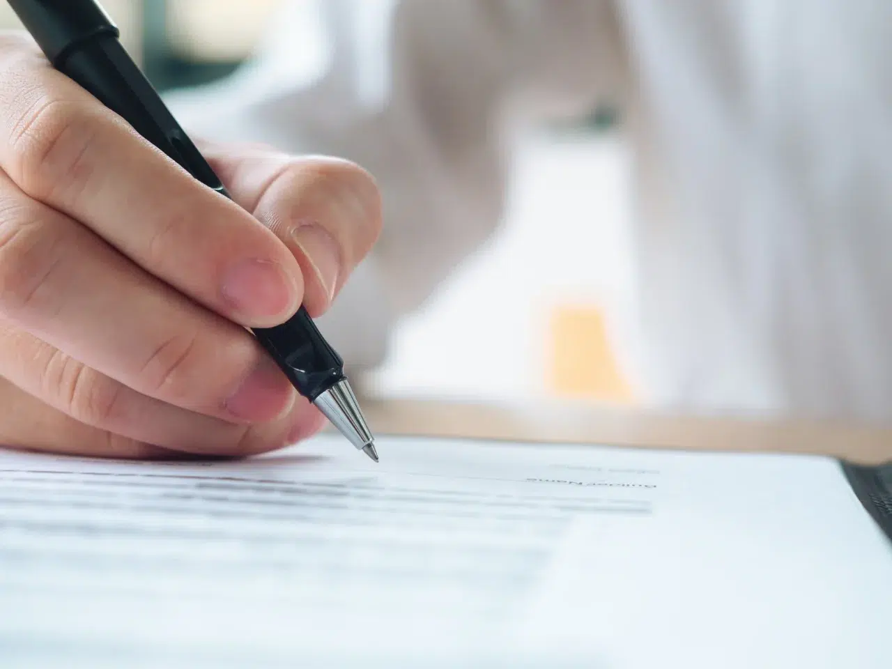 A close-up of a person’s hand holding a black pen and signing or writing on a document with printed text. The person is wearing a white shirt.