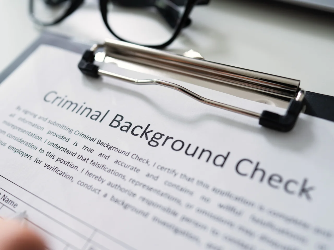 A close-up of a clipboard holding a document titled Criminal Background Check, with part of the text visible and a pair of eyeglasses blurred in the background.