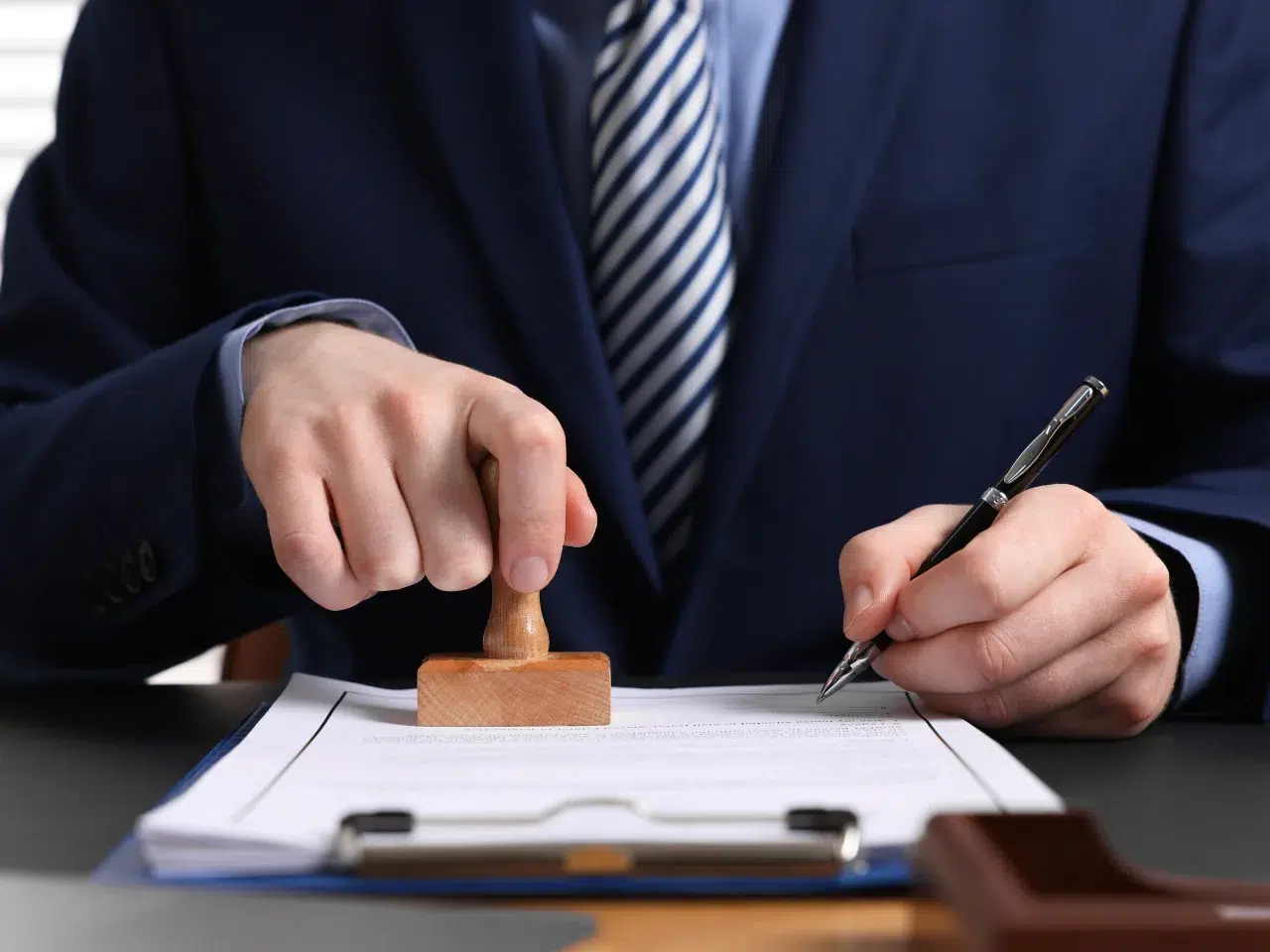 A person in a suit presses a wooden stamp onto a document while holding a pen, suggesting official approval or authorization of paperwork.