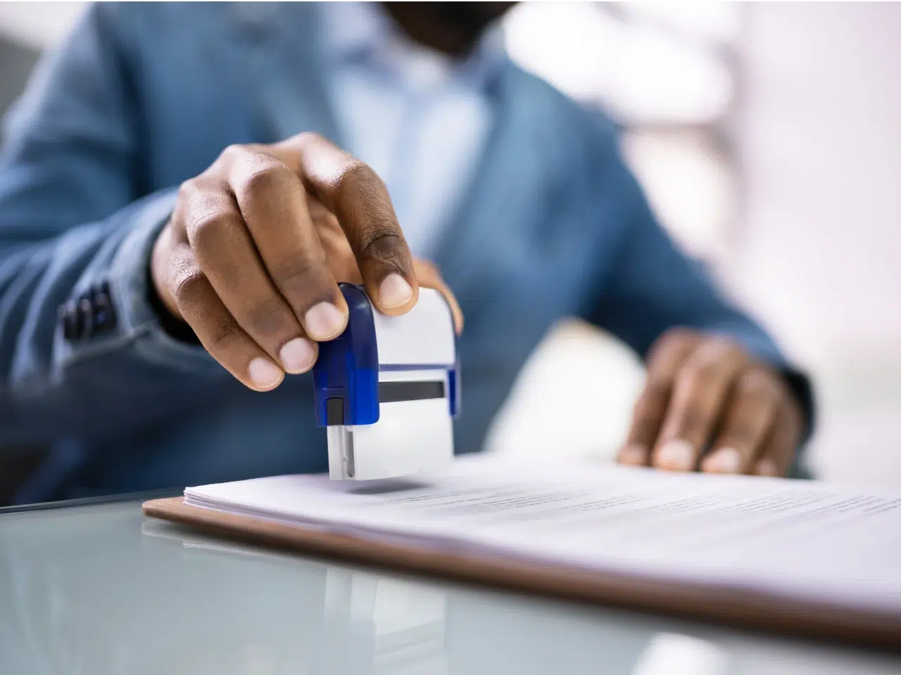 A person in a blue suit uses a stamp to mark a document on a clipboard, seated at a glass desk in an office setting.