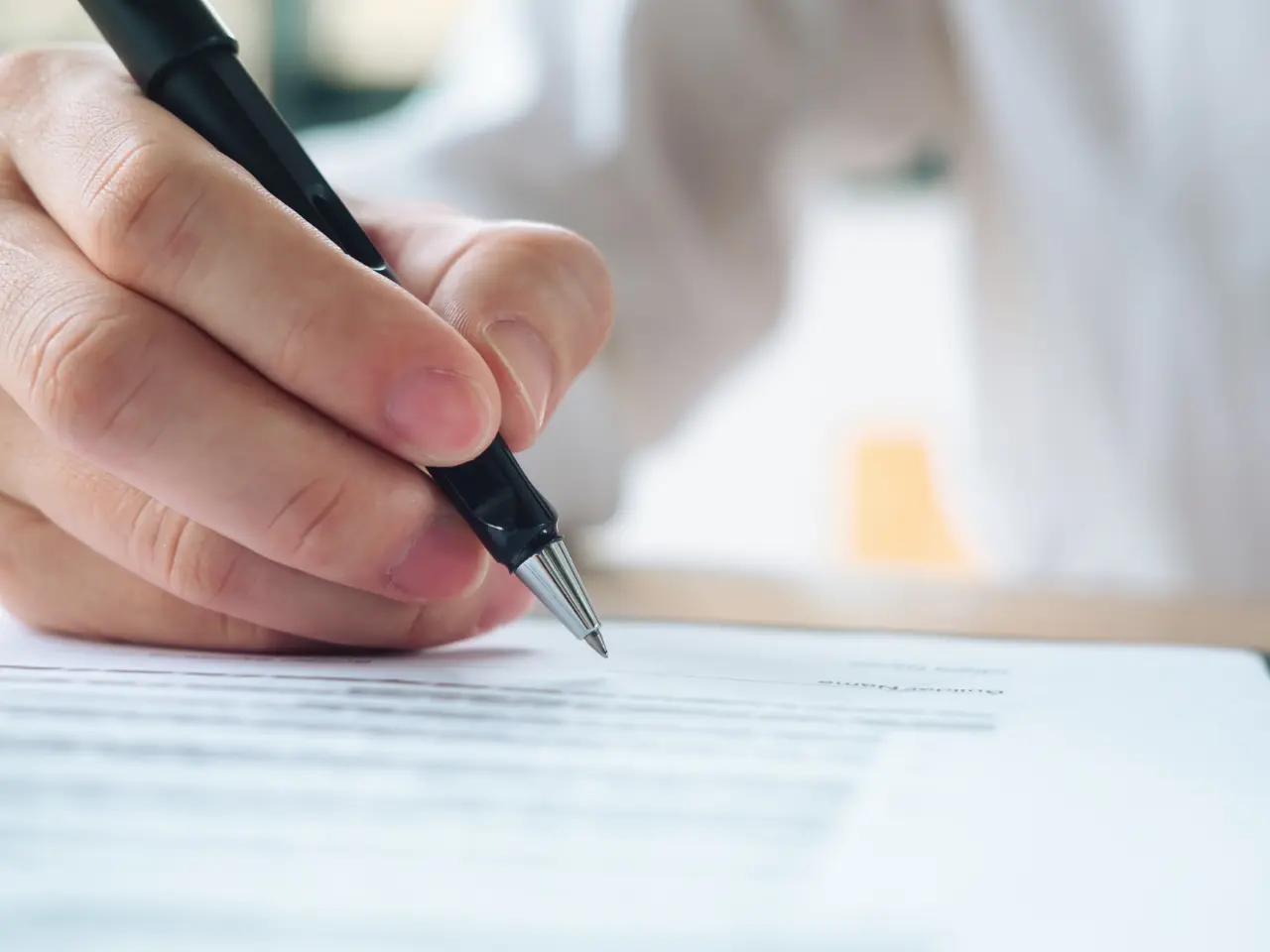 A close-up of a hand holding a pen and writing on a document, with the text on the paper blurred and a person in a light shirt visible in the background.