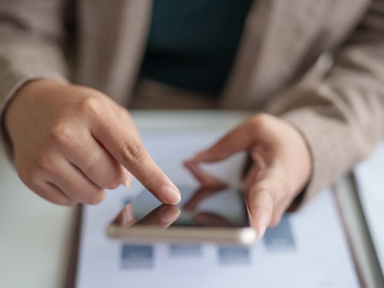 Close-up of a person’s hands using a smartphone, with one hand holding the phone and the other hand touching the screen. A blurred document is visible on the table in the background.
