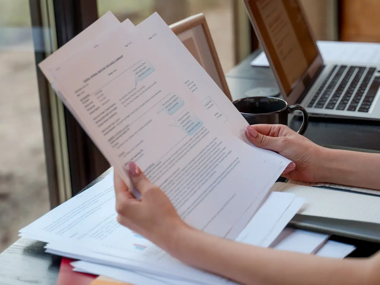 A person holding and reviewing printed documents at a desk with a laptop, a notebook, and a coffee mug nearby. Natural light comes through a window in the background.
