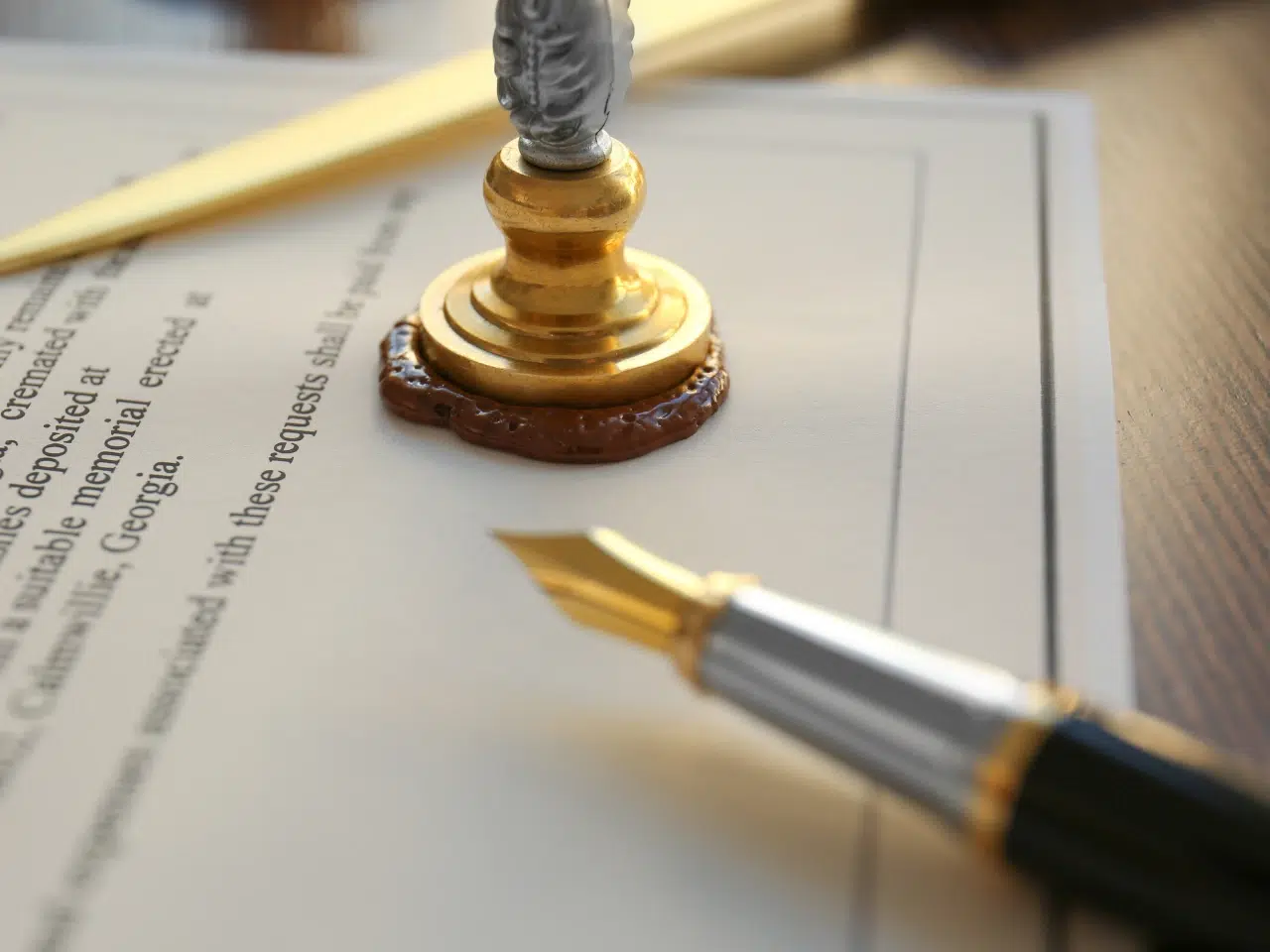 A close-up of a document being sealed with brown wax using a brass seal stamp, with a fountain pen and a letter opener nearby on a wooden surface.