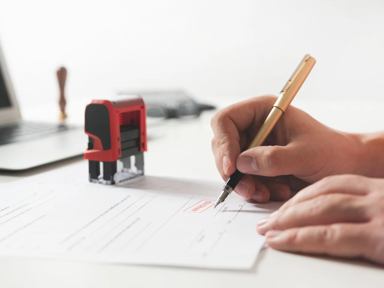 Close-up of a person signing a document with a gold pen next to a red and black approved stamp on a desk, with a laptop visible in the background.