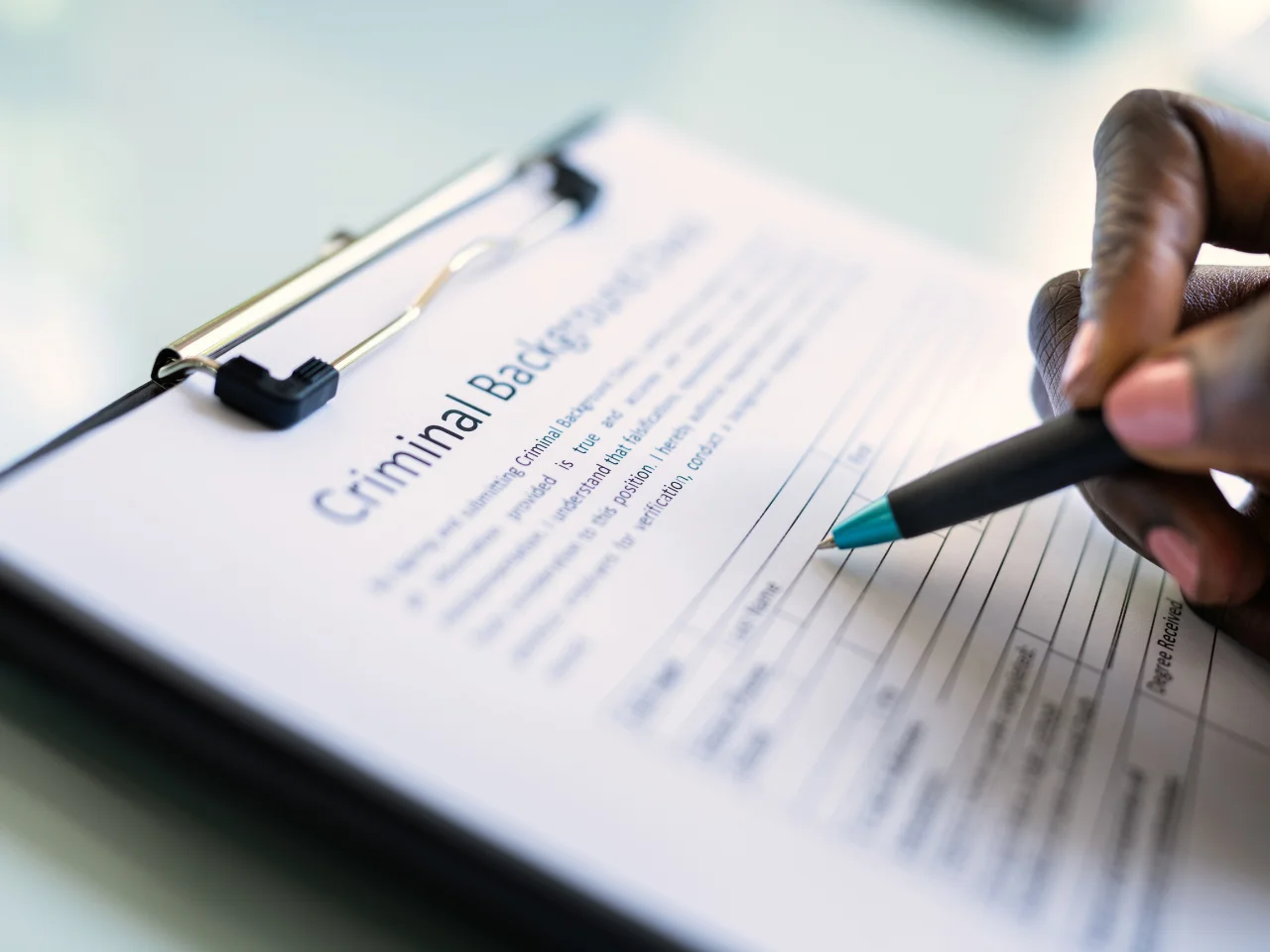 A close-up of a person filling out a criminal background check form on a clipboard with a turquoise pen. The form has blank spaces for personal information.