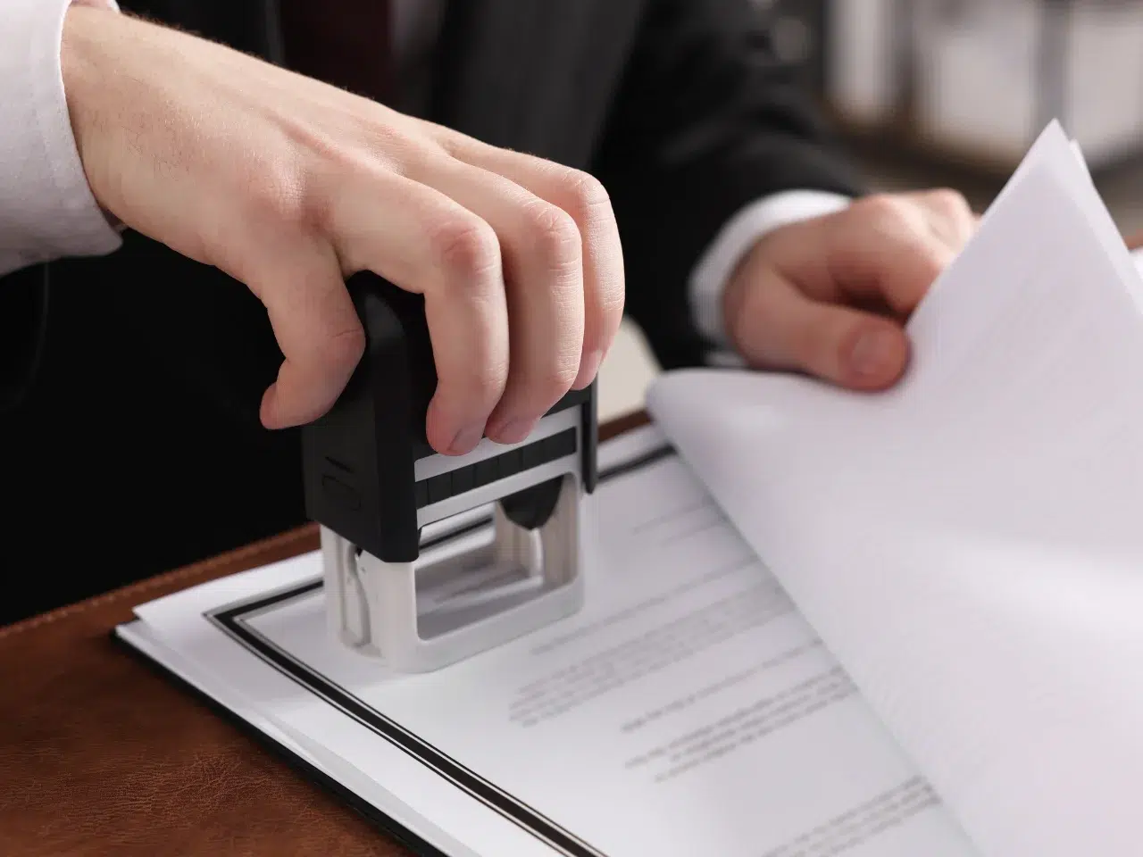 A person in a suit stamps a document on a desk while holding a stack of papers in the other hand, indicating the approval or authorization of official paperwork.