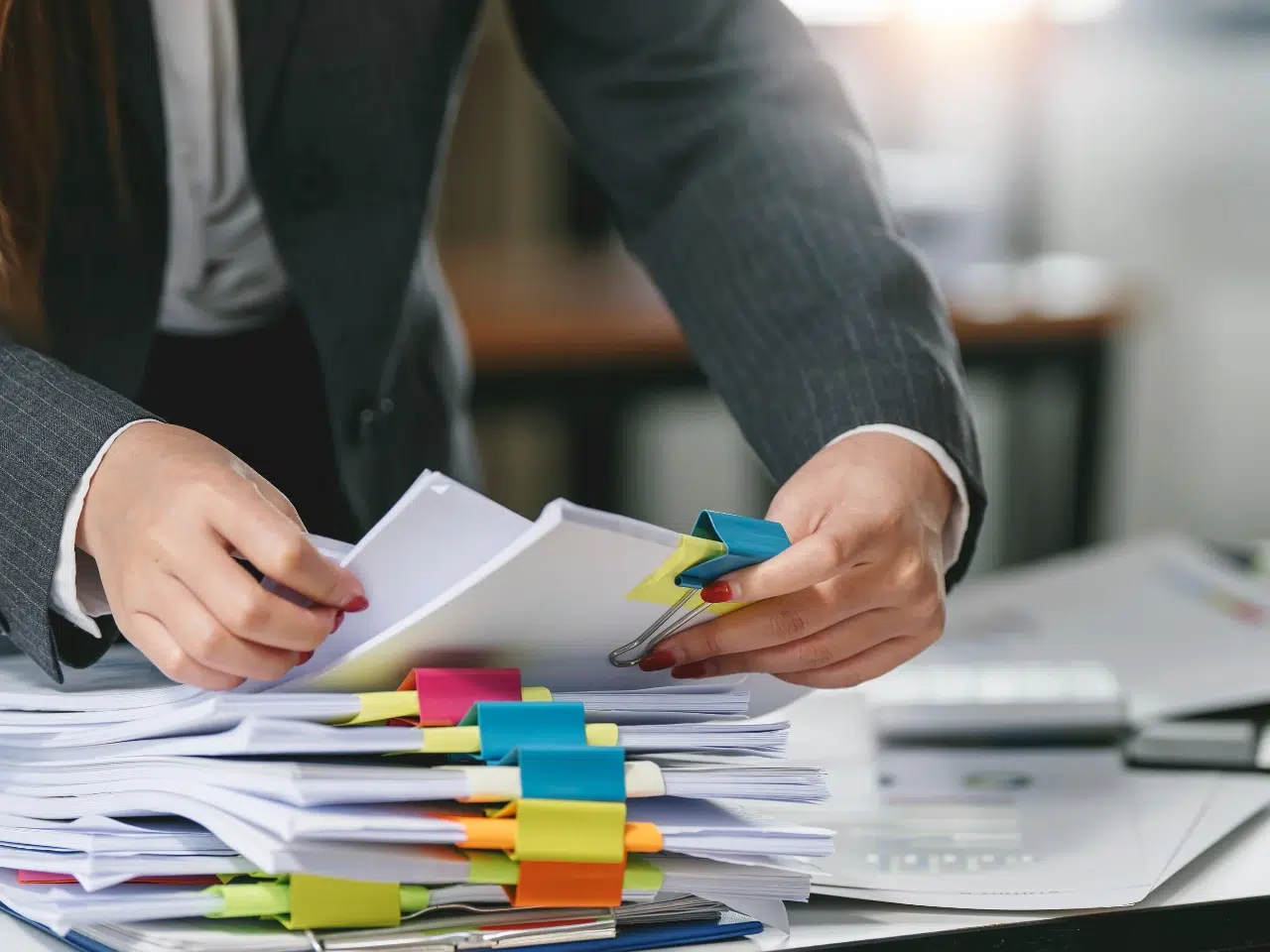 A person in a business suit organizes a large stack of documents on a desk, using colorful binder clips and sticky notes to separate sections. The background is blurred, suggesting an office setting.