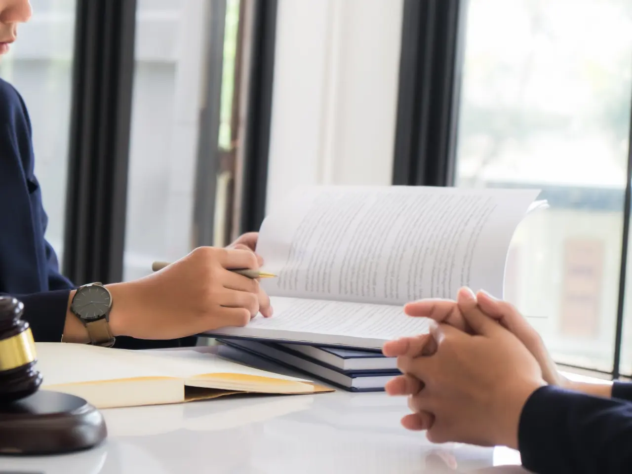 Two people sit across a desk from each other, one holding a pen and open book, with a gavel and more documents nearby, suggesting a legal or professional discussion in an office setting.