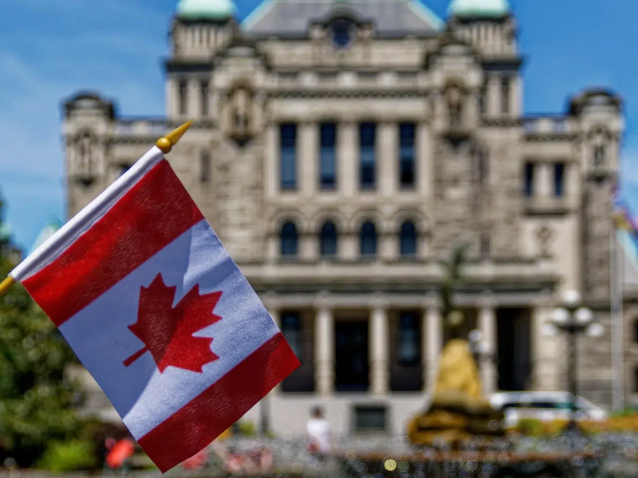 A small Canadian flag waves in the foreground with a historic stone building, possibly a government or parliamentary building, blurred in the background on a sunny day.