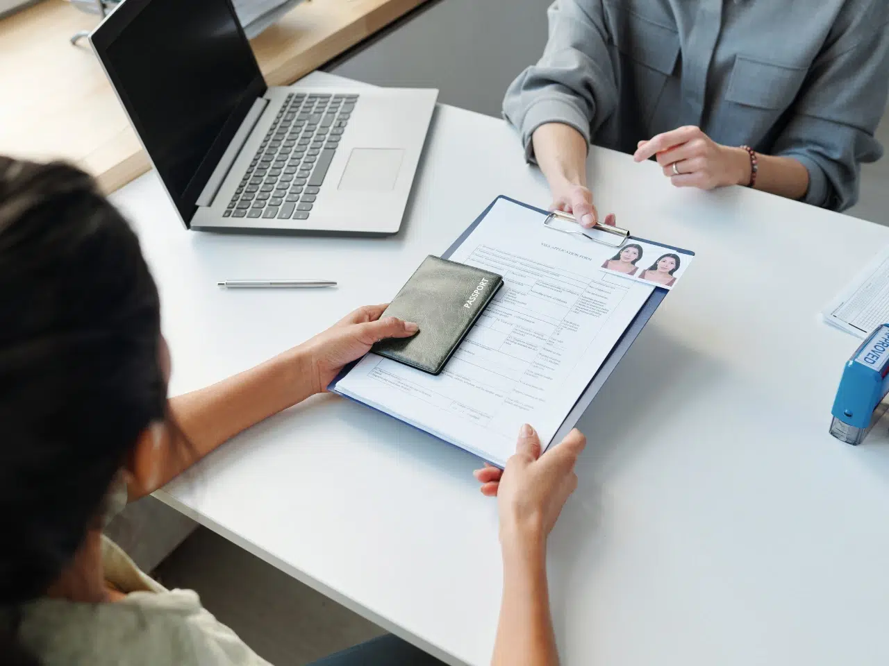 Two people sit at a desk with a laptop. One hands over a clipboard with a form and photos attached, along with an open passport, while the other points at the document.