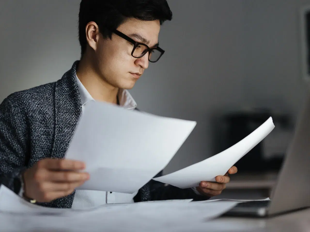 A person wearing glasses and a blazer is sitting at a desk, looking at papers in both hands, with a laptop and additional documents in front of them. The setting appears to be an office or workspace.