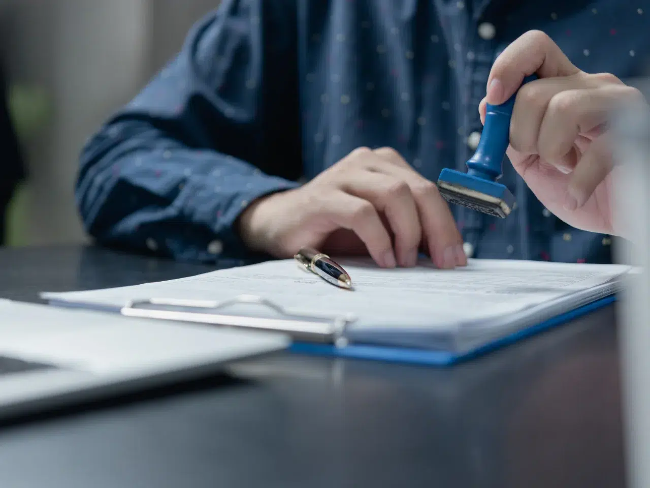 A person in a blue shirt stamps a document on a desk, with a pen and an open folder nearby.