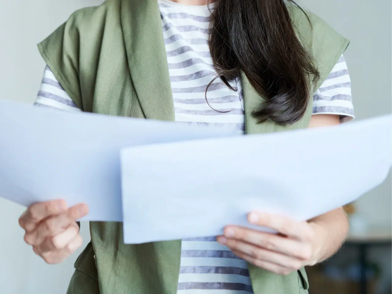 A person wearing a green vest over a striped shirt is holding and looking at two sheets of paper. Their face is not visible in the image.