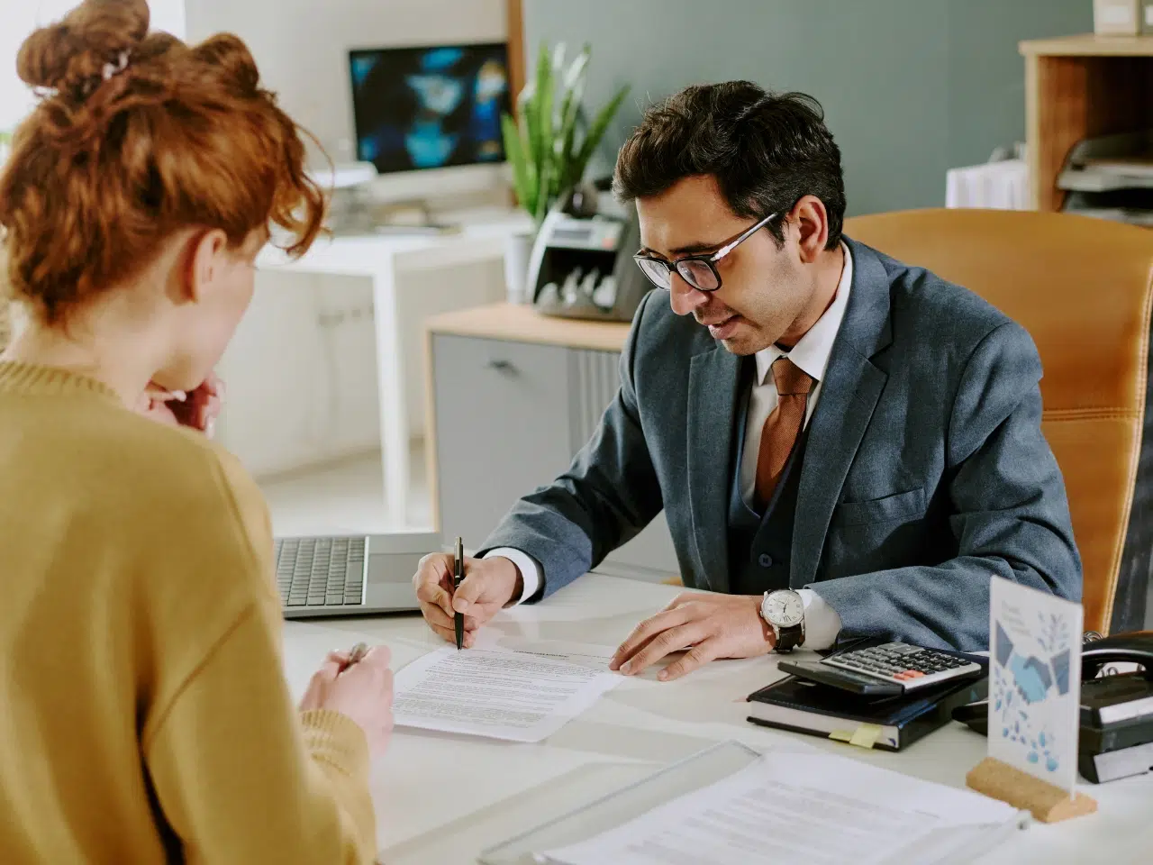 A man in a suit sits at a desk, reviewing and signing documents with a woman across from him in an office setting; a laptop, calculator, and papers are on the desk.