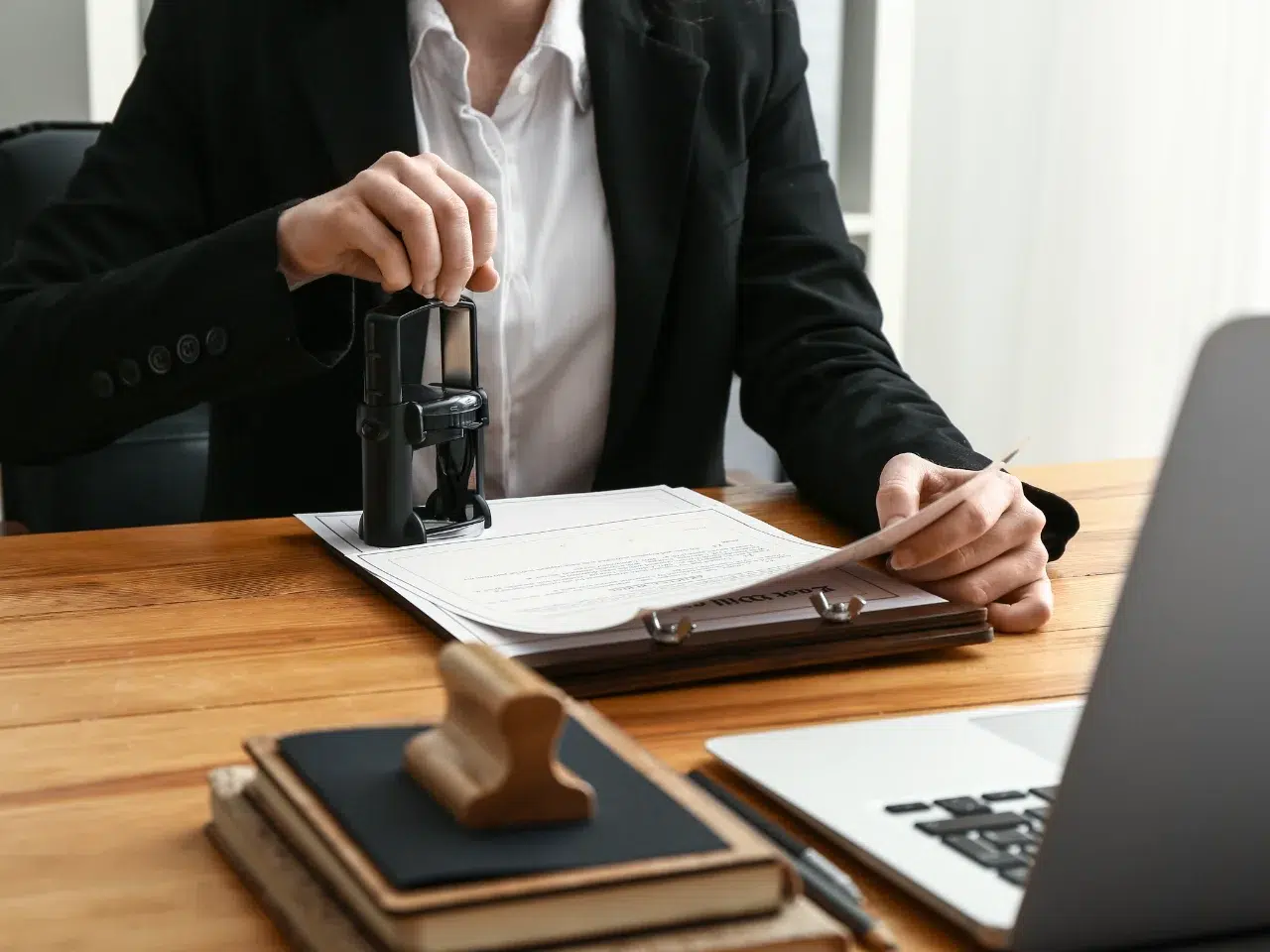 A person in a suit sits at a wooden desk, stamping a document on a clipboard. A laptop, a wooden stamp, and notebooks are also on the desk.
