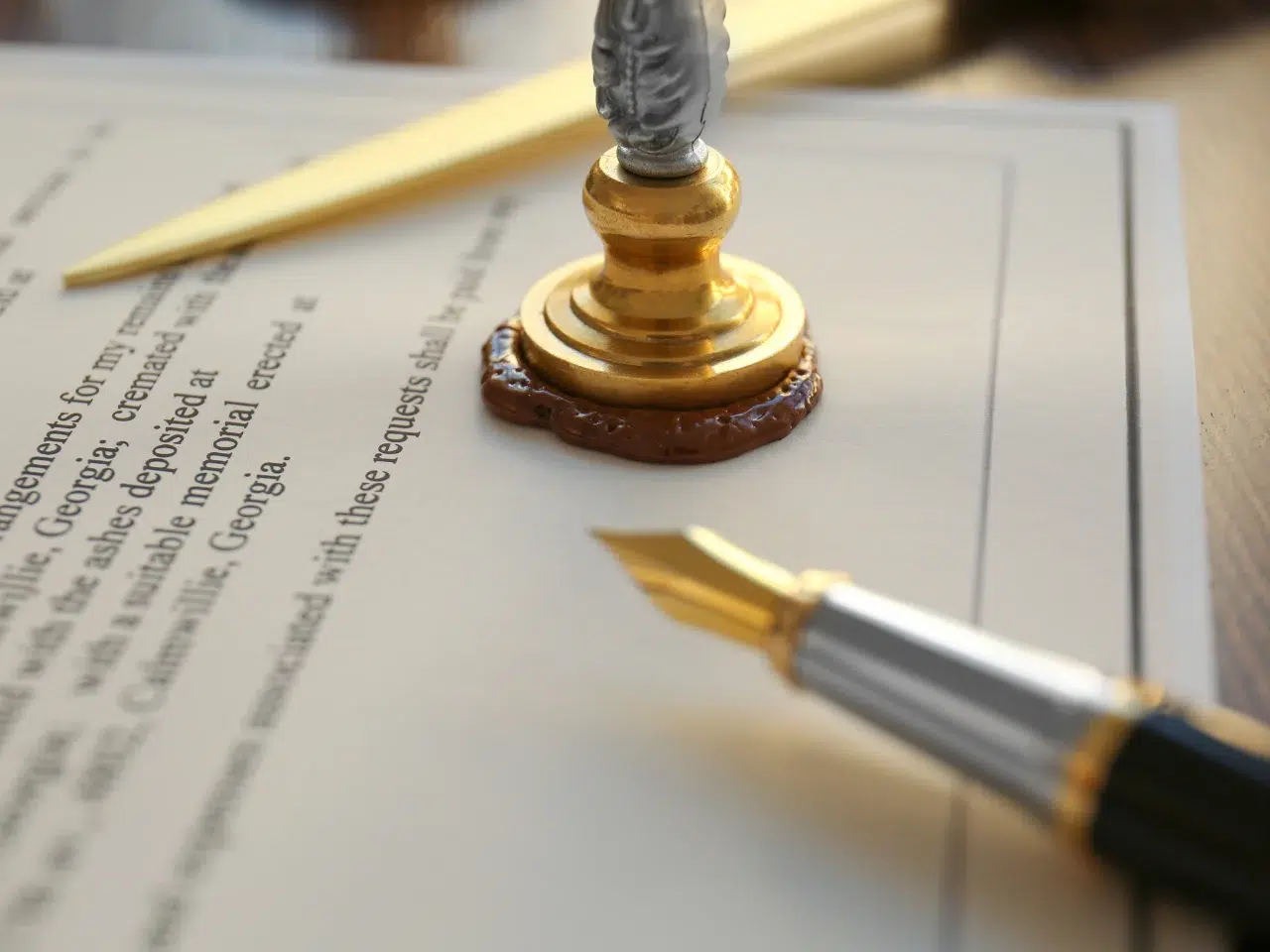 A close-up of a fountain pen, a sealed wax stamp, and a gold letter opener resting on a formal document. The wax seal is brown and the pen has a gold nib. The text on the document is partially visible.