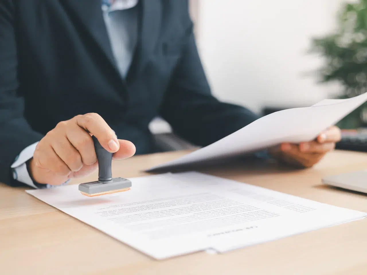 A person in a suit stamps a document on a desk while holding papers in their other hand, suggesting approval or official authorization in a business or legal setting.
