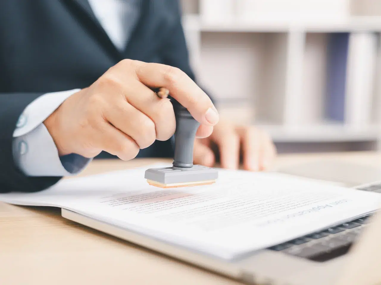 A person in a suit presses a rubber stamp onto a document placed on a laptop, suggesting the approval or authorization of paperwork in an office setting.