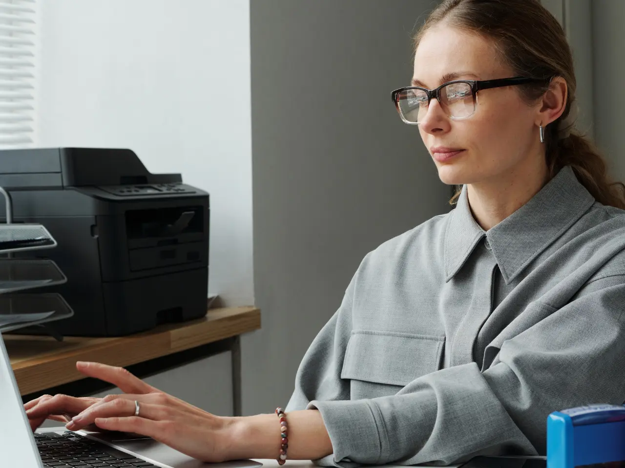 A woman wearing glasses and a gray shirt types on a laptop at a desk. A printer and file organizer are visible in the background. She appears focused on her work.