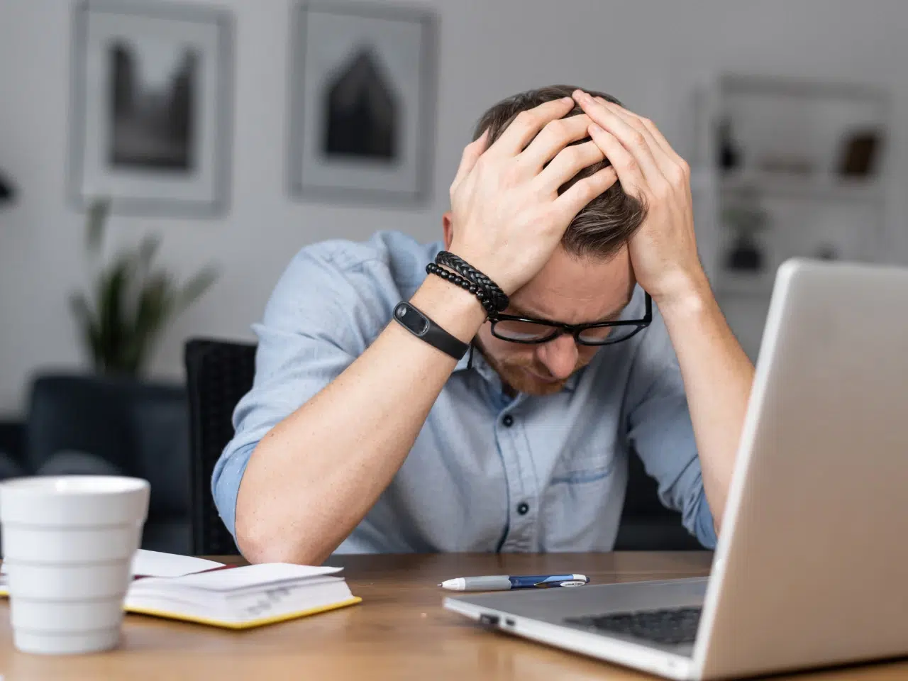 A man wearing glasses sits at a desk with his head in his hands, looking stressed. There is a laptop, notebook, pen, and coffee cup on the desk. The background shows blurred wall art and a plant.