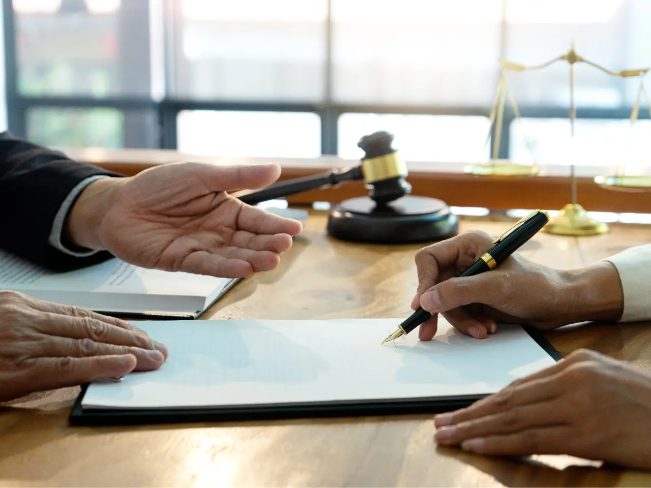 Two people sit at a desk; one gestures while the other signs a document. A judges gavel and scales of justice are visible on the table, suggesting a legal or official setting.