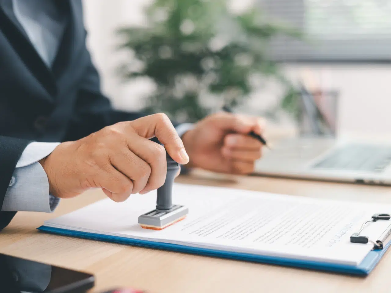 A person in business attire stamps a document on a desk, with a pen in the other hand. A laptop, phone, and clipboard are visible nearby, and a blurred plant is in the background.