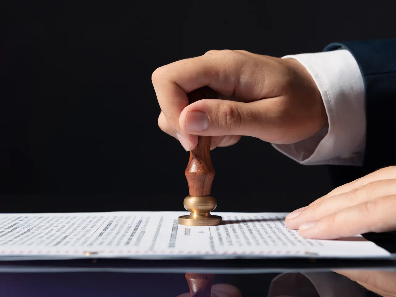 A person’s hand presses a wooden stamp onto a document with printed text, against a black background.